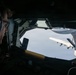 A boom operator aboard a tanker aircraft is observed monitoring an aerial refueling operation from the rear boom operator station. Through the observation window, a large multi-engine aircraft (appearing to be a C-130 or similar transport) is visible receiving or approaching for fuel transfer via a refueling boom. The interior is dimly lit, contrasting with the bright sky visible through the window.