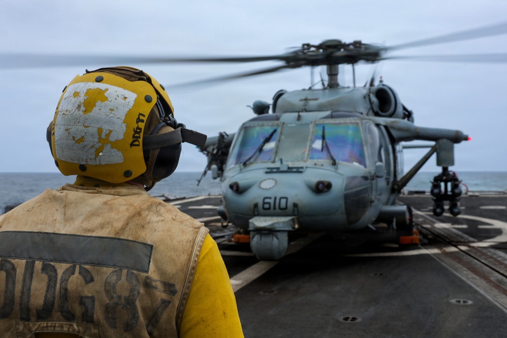 A naval flight deck crew member in yellow safety gear and helmet stands on a ship's flight deck observing a military helicopter numbered '610' with rotors spinning. The scene appears to take place at sea under overcast skies. The crew member's helmet and vest are marked with 'DDG-77', identifying the vessel as a U.S. Navy Arleigh Burke-class guided-missile destroyer.