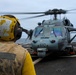 A naval flight deck crew member in yellow safety gear and helmet stands on a ship's flight deck observing a military helicopter numbered '610' with rotors spinning. The scene appears to take place at sea under overcast skies. The crew member's helmet and vest are marked with 'DDG-77', identifying the vessel as a U.S. Navy Arleigh Burke-class guided-missile destroyer.