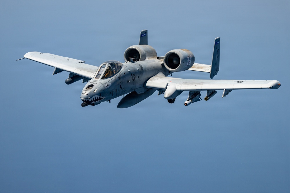 A U.S. Air Force A-10 Thunderbolt II (Warthog) attack aircraft is photographed in flight against a clear blue sky. The aircraft displays distinctive shark mouth nose art and carries visible ordnance on its wing pylons. The image is a high-quality aerial photograph taken in close proximity, likely from another aircraft.