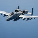 A U.S. Air Force A-10 Thunderbolt II (Warthog) attack aircraft is photographed in flight against a clear blue sky. The aircraft displays distinctive shark mouth nose art and carries visible ordnance on its wing pylons. The image is a high-quality aerial photograph taken in close proximity, likely from another aircraft.
