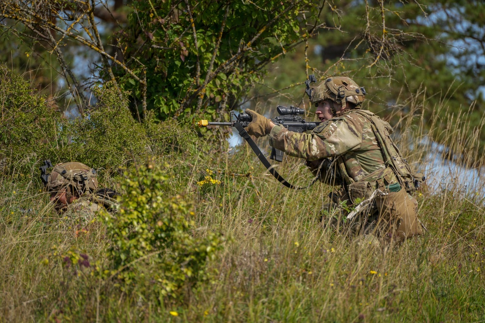Two soldiers in MultiCam/OCP camouflage uniforms are conducting what appears to be a military training exercise in a grassy, wooded field environment. The standing soldier is in a kneeling firing position aiming a rifle fitted with a yellow blank-firing adapter (BFA) and optical scope, while a second soldier is prone in the foreground. The presence of the yellow BFA on the muzzle and smoke in the background strongly indicates this is a live-fire training exercise rather than a combat situation.