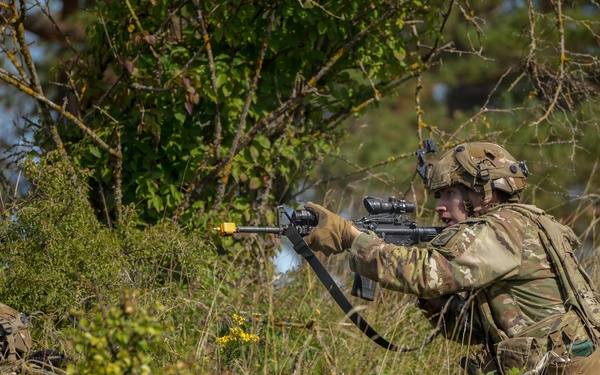 Two soldiers in MultiCam/OCP camouflage uniforms are conducting what appears to be a military training exercise in a grassy, wooded field environment. The standing soldier is in a kneeling firing position aiming a rifle fitted with a yellow blank-firing adapter (BFA) and optical scope, while a second soldier is prone in the foreground. The presence of the yellow BFA on the muzzle and smoke in the background strongly indicates this is a live-fire training exercise rather than a combat situation.