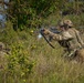 Two soldiers in MultiCam/OCP camouflage uniforms are conducting what appears to be a military training exercise in a grassy, wooded field environment. The standing soldier is in a kneeling firing position aiming a rifle fitted with a yellow blank-firing adapter (BFA) and optical scope, while a second soldier is prone in the foreground. The presence of the yellow BFA on the muzzle and smoke in the background strongly indicates this is a live-fire training exercise rather than a combat situation.