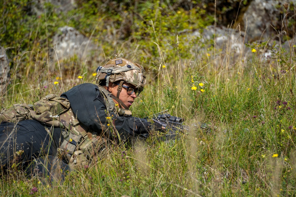 A soldier in full tactical gear is lying prone in a grassy field with wildflowers, appearing to be in a low-profile observation or defensive position. The individual is equipped with a combat helmet, tactical vest, backpack, gloves, and protective eyewear, and is holding a firearm. The setting appears to be a field training exercise or operational scenario in a natural outdoor environment.