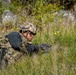 A soldier in full tactical gear is lying prone in a grassy field with wildflowers, appearing to be in a low-profile observation or defensive position. The individual is equipped with a combat helmet, tactical vest, backpack, gloves, and protective eyewear, and is holding a firearm. The setting appears to be a field training exercise or operational scenario in a natural outdoor environment.