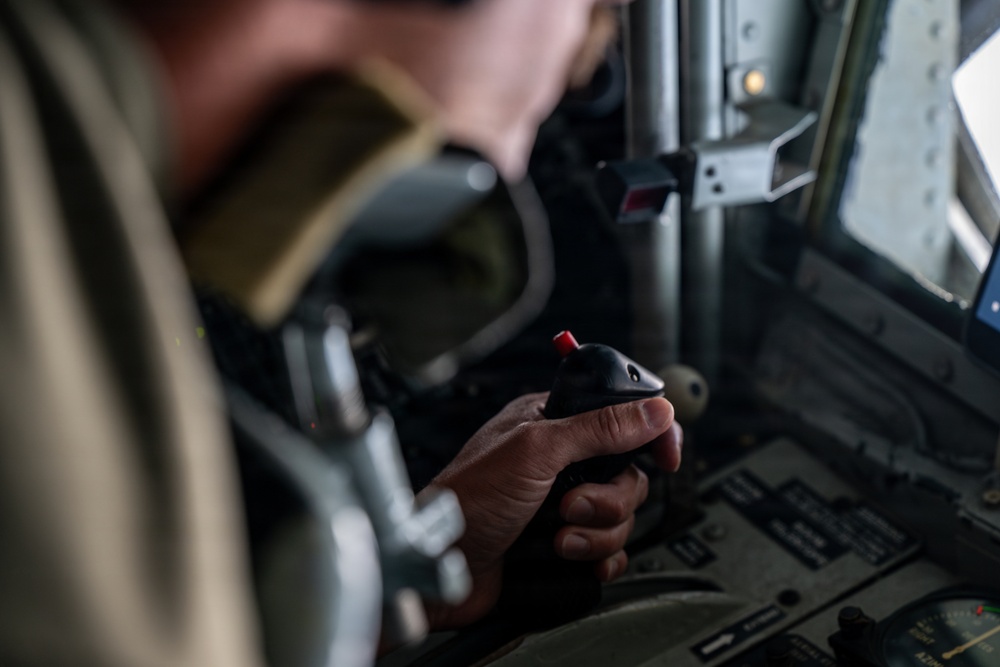 A person in what appears to be a flight suit or military uniform is gripping a control stick (joystick) inside a military aircraft cockpit. The joystick features a prominent red button on top and is mounted on a control column. The cockpit background shows instrument panels and a side window with natural light.
