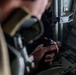 A person in what appears to be a flight suit or military uniform is gripping a control stick (joystick) inside a military aircraft cockpit. The joystick features a prominent red button on top and is mounted on a control column. The cockpit background shows instrument panels and a side window with natural light.
