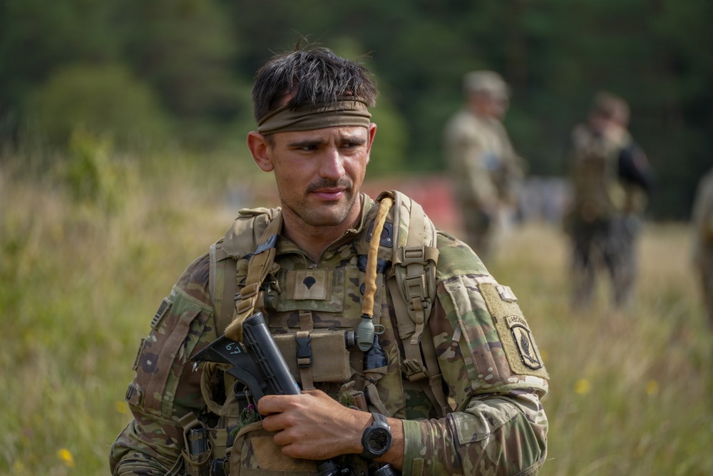 A male soldier in MultiCam/OCP camouflage uniform is photographed in a field environment, holding what appears to be a shotgun or grenade launcher. He wears a tactical vest, headband, and has an 'Airborne' tab patch visible on his left shoulder. Additional soldiers are visible in the blurred background.