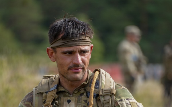 A male soldier in MultiCam/OCP camouflage uniform is photographed in a field environment, holding what appears to be a shotgun or grenade launcher. He wears a tactical vest, headband, and has an 'Airborne' tab patch visible on his left shoulder. Additional soldiers are visible in the blurred background.