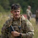 A male soldier in MultiCam/OCP camouflage uniform is photographed in a field environment, holding what appears to be a shotgun or grenade launcher. He wears a tactical vest, headband, and has an 'Airborne' tab patch visible on his left shoulder. Additional soldiers are visible in the blurred background.