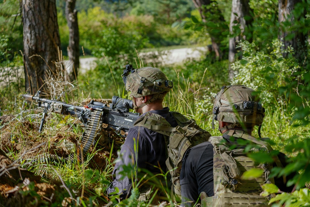 Two military personnel in full tactical gear are positioned in a wooded area, with one operating what appears to be a belt-fed machine gun mounted on a bipod. Both individuals are wearing camouflage uniforms, tactical vests, and helmets equipped with night-vision device mounts. The setting appears to be a training exercise or field operation in a forested environment.