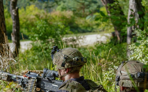 Two military personnel in full tactical gear are positioned in a wooded area, with one operating what appears to be a belt-fed machine gun mounted on a bipod. Both individuals are wearing camouflage uniforms, tactical vests, and helmets equipped with night-vision device mounts. The setting appears to be a training exercise or field operation in a forested environment.