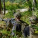 Two military personnel in full tactical gear are positioned in a wooded area, with one operating what appears to be a belt-fed machine gun mounted on a bipod. Both individuals are wearing camouflage uniforms, tactical vests, and helmets equipped with night-vision device mounts. The setting appears to be a training exercise or field operation in a forested environment.