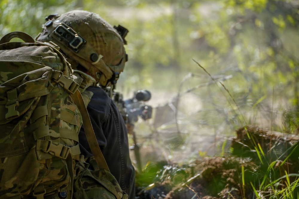 A soldier in full combat gear is positioned in a wooded area, aiming a rifle equipped with an optical scope. The individual is wearing multicam camouflage, a tactical helmet with mounted equipment, and carrying a large rucksack. Smoke or dust is visible near the muzzle area, suggesting recent or ongoing weapons discharge.