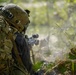 A soldier in full combat gear is positioned in a wooded area, aiming a rifle equipped with an optical scope. The individual is wearing multicam camouflage, a tactical helmet with mounted equipment, and carrying a large rucksack. Smoke or dust is visible near the muzzle area, suggesting recent or ongoing weapons discharge.