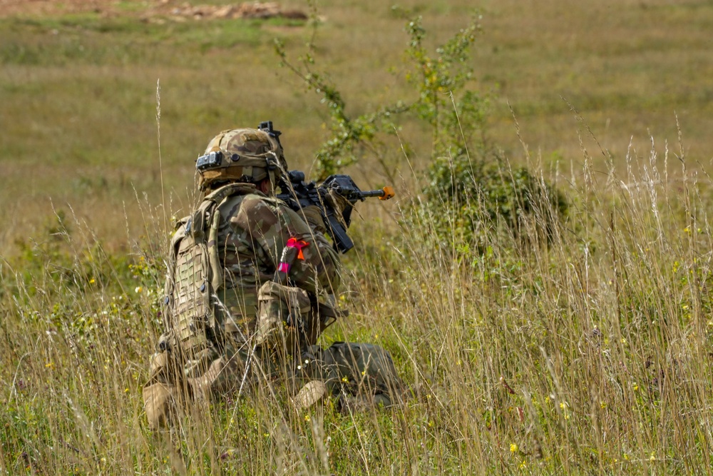 A soldier in full combat gear is kneeling in a grassy field, aiming a rifle forward. The soldier is wearing MultiCam/MTP camouflage pattern uniform with a tactical backpack and helmet. Bright pink/red ribbon markers and an orange muzzle cap on the weapon suggest this is a training exercise rather than live combat.