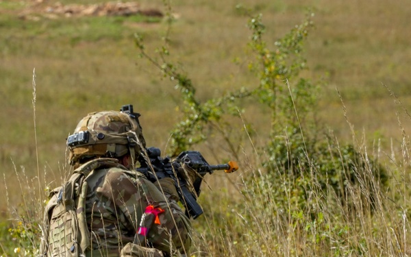 A soldier in full combat gear is kneeling in a grassy field, aiming a rifle forward. The soldier is wearing MultiCam/MTP camouflage pattern uniform with a tactical backpack and helmet. Bright pink/red ribbon markers and an orange muzzle cap on the weapon suggest this is a training exercise rather than live combat.