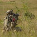 A soldier in full combat gear is kneeling in a grassy field, aiming a rifle forward. The soldier is wearing MultiCam/MTP camouflage pattern uniform with a tactical backpack and helmet. Bright pink/red ribbon markers and an orange muzzle cap on the weapon suggest this is a training exercise rather than live combat.