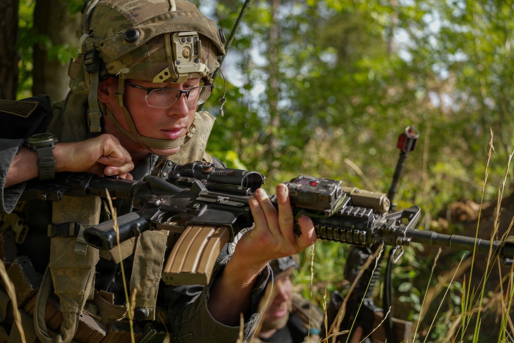 A soldier in full combat gear is holding and inspecting an assault rifle in a wooded outdoor environment. The soldier is wearing a tactical helmet with mounted equipment, ballistic eyewear, body armor, and a communications headset. A second partially visible individual is present in the background.