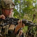A soldier in full combat gear is holding and inspecting an assault rifle in a wooded outdoor environment. The soldier is wearing a tactical helmet with mounted equipment, ballistic eyewear, body armor, and a communications headset. A second partially visible individual is present in the background.