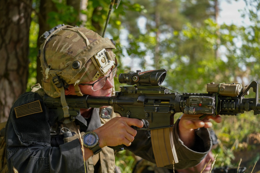 A U.S. Army soldier in full combat gear is aiming a rifle equipped with multiple optical attachments in a wooded outdoor environment. The soldier wears a multicam helmet with mounted equipment, ballistic glasses, and a uniform bearing a U.S. flag patch. The image appears to be taken during a training exercise or field operation.