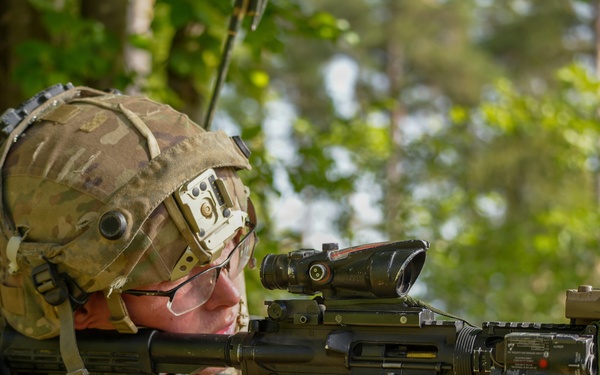 A U.S. Army soldier in full combat gear is aiming a rifle equipped with multiple optical attachments in a wooded outdoor environment. The soldier wears a multicam helmet with mounted equipment, ballistic glasses, and a uniform bearing a U.S. flag patch. The image appears to be taken during a training exercise or field operation.