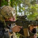 A U.S. Army soldier in full combat gear is aiming a rifle equipped with multiple optical attachments in a wooded outdoor environment. The soldier wears a multicam helmet with mounted equipment, ballistic glasses, and a uniform bearing a U.S. flag patch. The image appears to be taken during a training exercise or field operation.