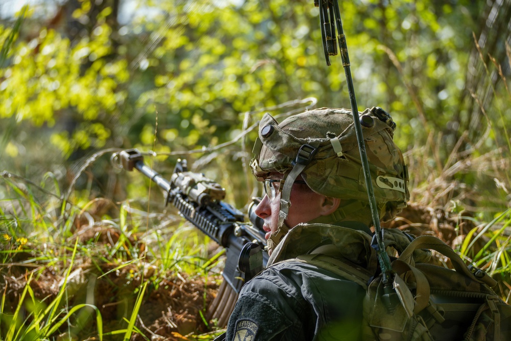 A soldier in full combat gear is positioned low in dense vegetation, aiming a rifle forward in what appears to be a field training exercise or tactical operation. The soldier is equipped with a camouflage helmet, tactical backpack, and a radio antenna extending upward. The image is captured in a sunlit outdoor environment with lush green foliage in the background.