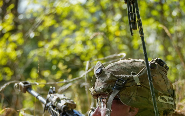 A soldier in full combat gear is positioned low in dense vegetation, aiming a rifle forward in what appears to be a field training exercise or tactical operation. The soldier is equipped with a camouflage helmet, tactical backpack, and a radio antenna extending upward. The image is captured in a sunlit outdoor environment with lush green foliage in the background.