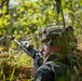 A soldier in full combat gear is positioned low in dense vegetation, aiming a rifle forward in what appears to be a field training exercise or tactical operation. The soldier is equipped with a camouflage helmet, tactical backpack, and a radio antenna extending upward. The image is captured in a sunlit outdoor environment with lush green foliage in the background.