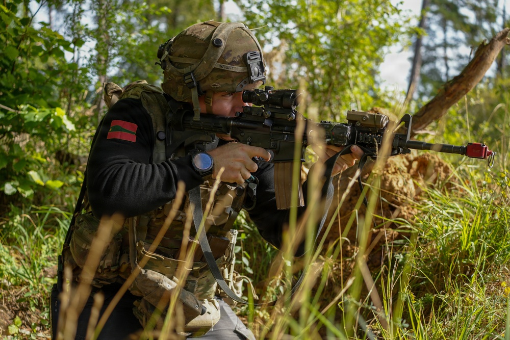 A soldier in tactical gear is crouching in a wooded area while aiming a rifle equipped with an optic sight. The individual is wearing a multicam helmet with a mounted device, a tactical vest over a black sweater, and has a name tape reading 'WRIGHT' visible on the helmet. The setting appears to be a training exercise, indicated by the red training barrel cap on the rifle muzzle.