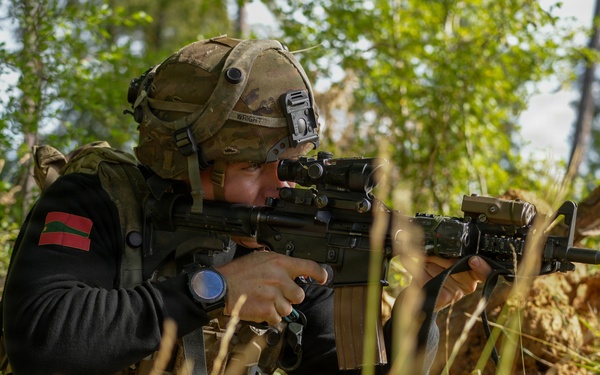 A soldier in tactical gear is crouching in a wooded area while aiming a rifle equipped with an optic sight. The individual is wearing a multicam helmet with a mounted device, a tactical vest over a black sweater, and has a name tape reading 'WRIGHT' visible on the helmet. The setting appears to be a training exercise, indicated by the red training barrel cap on the rifle muzzle.
