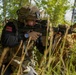 A soldier in tactical gear is crouching in a wooded area while aiming a rifle equipped with an optic sight. The individual is wearing a multicam helmet with a mounted device, a tactical vest over a black sweater, and has a name tape reading 'WRIGHT' visible on the helmet. The setting appears to be a training exercise, indicated by the red training barrel cap on the rifle muzzle.