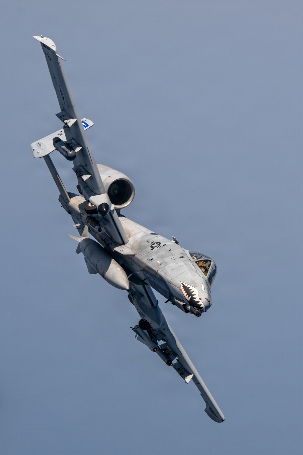 A U.S. Air Force A-10 Thunderbolt II (Warthog) is captured in a steep banking maneuver against a gray-blue sky. The aircraft features a distinctive shark mouth nose art painted on the fuselage and is carrying multiple underwing stores. The image is taken from below and slightly to the side, providing a dramatic view of the aircraft's underside and twin rear-mounted turbofan engines.