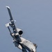 A U.S. Air Force A-10 Thunderbolt II (Warthog) is captured in a steep banking maneuver against a gray-blue sky. The aircraft features a distinctive shark mouth nose art painted on the fuselage and is carrying multiple underwing stores. The image is taken from below and slightly to the side, providing a dramatic view of the aircraft's underside and twin rear-mounted turbofan engines.