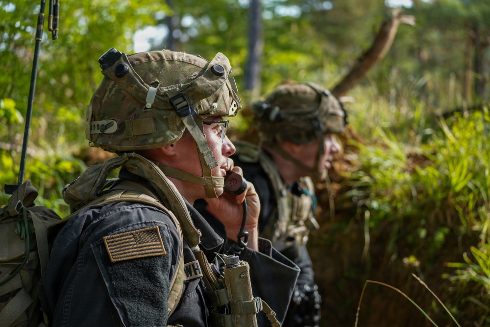 Two U.S. Army soldiers in full combat gear are positioned in a wooded, vegetated area. The foreground soldier is using a radio handset to communicate while wearing multicam camouflage helmet and body armor with a visible U.S. flag patch. A second soldier is visible in the background, similarly equipped.