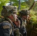 Two U.S. Army soldiers in full combat gear are positioned in a wooded, vegetated area. The foreground soldier is using a radio handset to communicate while wearing multicam camouflage helmet and body armor with a visible U.S. flag patch. A second soldier is visible in the background, similarly equipped.