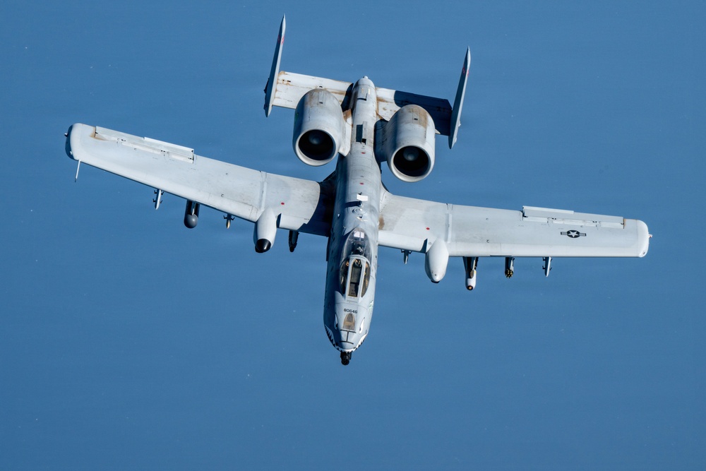 A U.S. Air Force A-10 Thunderbolt II (Warthog) attack aircraft is photographed in flight from a slightly elevated angle, showing a clear top-down perspective. The aircraft is painted in a light gray scheme and carries visible ordnance on its wing hardpoints. The image was taken in clear daylight conditions against a bright blue sky.