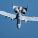 A U.S. Air Force A-10 Thunderbolt II (Warthog) attack aircraft is photographed in flight from a slightly elevated angle, showing a clear top-down perspective. The aircraft is painted in a light gray scheme and carries visible ordnance on its wing hardpoints. The image was taken in clear daylight conditions against a bright blue sky.