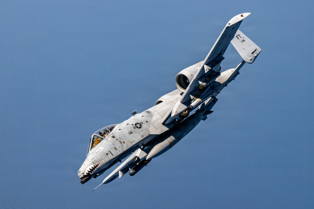 A U.S. Air Force A-10 Thunderbolt II (Warthog) attack aircraft is captured in a steep banking maneuver against a clear blue sky, with what appears to be a body of water visible below. The aircraft displays the iconic shark mouth nose art and carries visible ordnance/weapons on its wing pylons. The image is a high-quality aerial photograph likely taken from another aircraft.