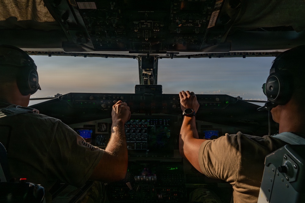 Two military aircrew members are seated in the cockpit of a large military transport aircraft, viewed from behind. Both pilots are wearing olive drab flight suits and aviation headsets, with hands on or near the controls. The cockpit features a complex instrument panel with multiple digital displays, and the view through the windscreen shows open sky at what appears to be dusk or dawn.