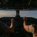 Two military aircrew members are seated in the cockpit of a large military transport aircraft, viewed from behind. Both pilots are wearing olive drab flight suits and aviation headsets, with hands on or near the controls. The cockpit features a complex instrument panel with multiple digital displays, and the view through the windscreen shows open sky at what appears to be dusk or dawn.