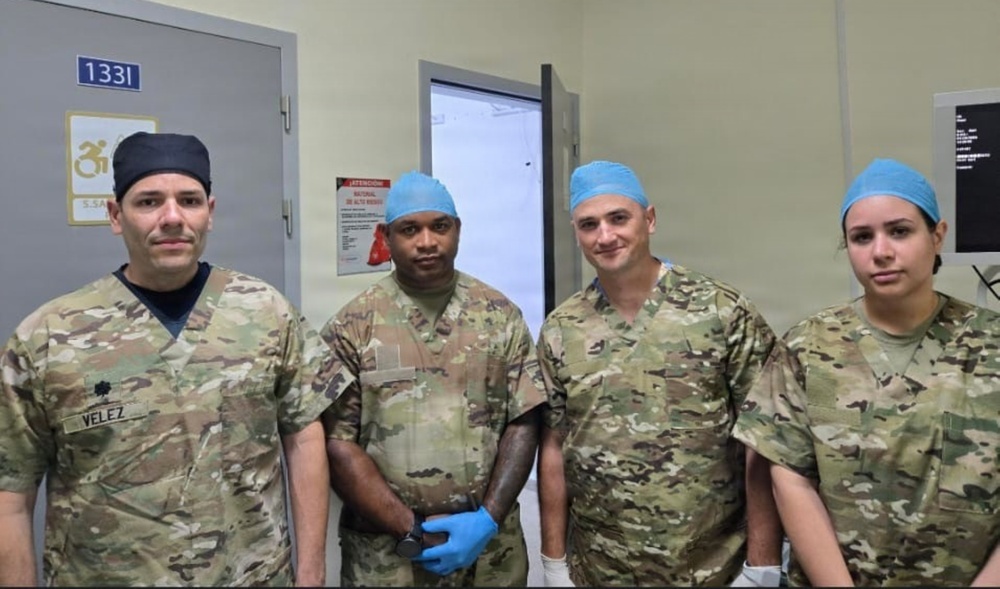 Four military medical personnel are posing together in what appears to be a hospital or medical facility corridor. They are wearing US Army OCP (Operational Camouflage Pattern) uniforms combined with surgical scrub caps, indicating they are military healthcare providers. One individual on the left has a name tape reading 'VELEZ' and wears a black scrub cap, while the other three wear blue surgical caps.
