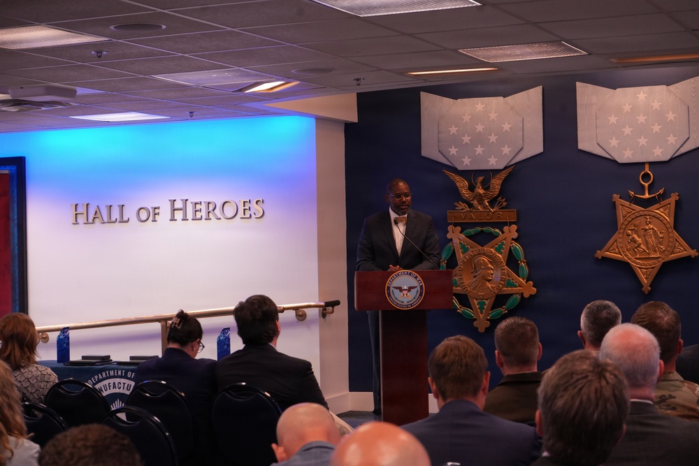 A speaker stands at a podium bearing the Department of Defense seal in the Pentagon's Hall of Heroes, addressing a seated audience. The room features large Medal of Honor emblems on a navy blue wall, including the Army and Navy versions of the medal. The event appears to be a formal ceremony or official presentation.