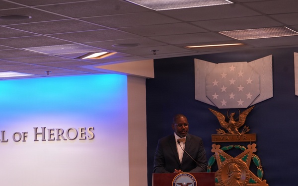A speaker stands at a podium bearing the Department of Defense seal in the Pentagon's Hall of Heroes, addressing a seated audience. The room features large Medal of Honor emblems on a navy blue wall, including the Army and Navy versions of the medal. The event appears to be a formal ceremony or official presentation.