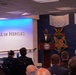 A speaker stands at a podium bearing the Department of Defense seal in the Pentagon's Hall of Heroes, addressing a seated audience. The room features large Medal of Honor emblems on a navy blue wall, including the Army and Navy versions of the medal. The event appears to be a formal ceremony or official presentation.