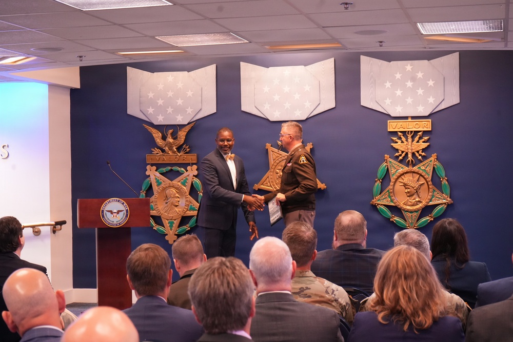 A formal ceremony is taking place at what appears to be the Pentagon or a Department of Defense facility, where a civilian in a suit is shaking hands with a uniformed Army soldier on a stage. The backdrop features large decorative Medal of Honor emblems with 'VALOR' inscriptions and star-spangled banners. An audience of military and civilian personnel is seated in the foreground observing the event.
