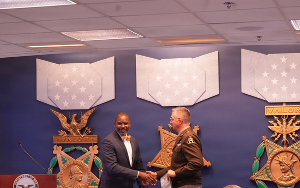 A formal ceremony is taking place at what appears to be the Pentagon or a Department of Defense facility, where a civilian in a suit is shaking hands with a uniformed Army soldier on a stage. The backdrop features large decorative Medal of Honor emblems with 'VALOR' inscriptions and star-spangled banners. An audience of military and civilian personnel is seated in the foreground observing the event.