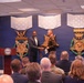 A formal ceremony is taking place at what appears to be the Pentagon or a Department of Defense facility, where a civilian in a suit is shaking hands with a uniformed Army soldier on a stage. The backdrop features large decorative Medal of Honor emblems with 'VALOR' inscriptions and star-spangled banners. An audience of military and civilian personnel is seated in the foreground observing the event.